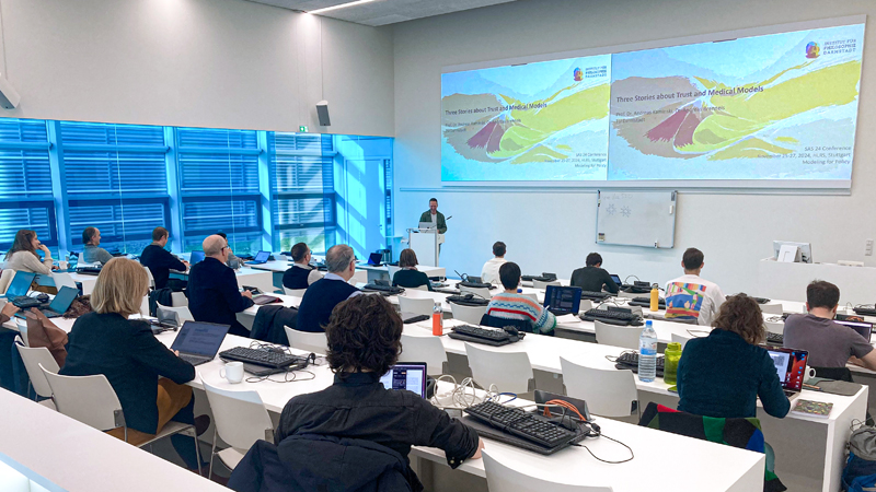 Foto des HLRS-Konferenzraums auf der SAS 24-Konferenz. Auf dem Bildschirm ist eine Folie mit dem Titel „Three Stories about Trust and Medical Models“ zu sehen.